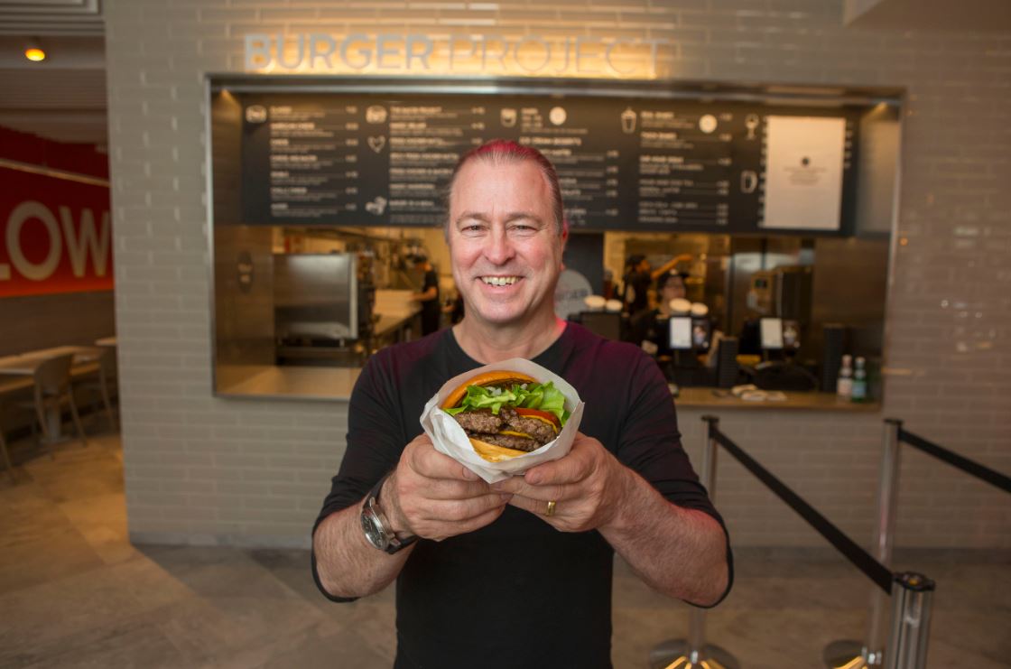 Neil Perry holding a burger and smiling at the opening of a new Burger Project.