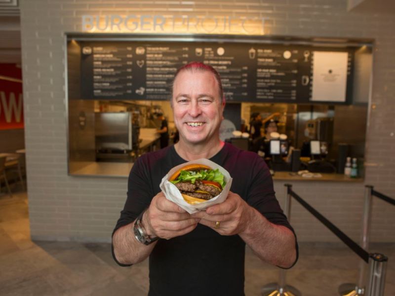 Neil Perry holding a burger and smiling at the opening of a new Burger Project.