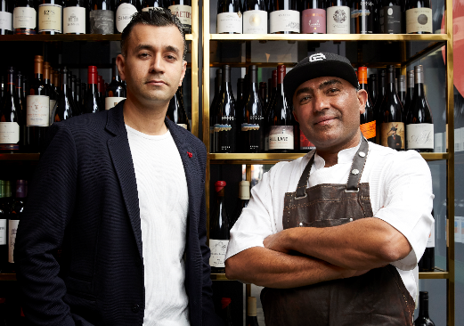 Dheeraj Bhatia and Jessi Singh looking into camera, standing in front of wine-filled shelves.